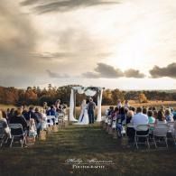 Outdoor wedding ceremony under a white arch at sunset, with guests seated in rows on both sides.