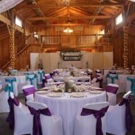 Banquet hall with round tables, white linens, and purple chair sashes under warm wooden beams