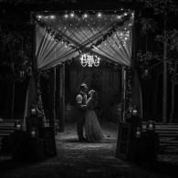 Black-and-white wedding couple dancing under a draped archway with chandelier lights at night