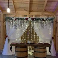 Rustic wedding reception setup with a decorated arch, twinkle lights, and a wooden table on barrel stands