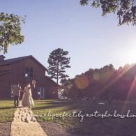 Two people walking on a stone path by a barn at sunset, with long shadows and pink lens flare