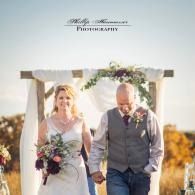 Bride and groom walking hand in hand under a floral arch outdoors at sunset