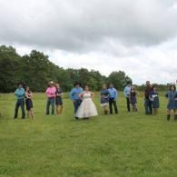 People standing in a grassy field outdoors, with one person in a white dress at center under a cloudy sky