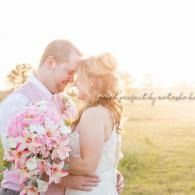 Couple embracing in a sunny field, holding a bouquet of pink flowers