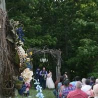 Outdoor wedding ceremony under a floral arch, with bride and officiant at the altar and guests seated outside.