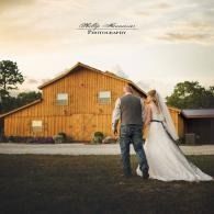 Bride and groom walking hand in hand toward a wooden barn at sunset