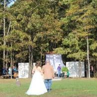 Bride and groom walk toward an outdoor wedding altar in a grassy clearing surrounded by trees.