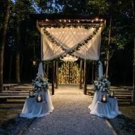 Outdoor wedding ceremony arch with white drapery, lanterns, and floral arrangements on a forest path at dusk