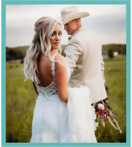 Bride and groom in a field, the bride looking back in a white dress and the groom in a hat holding flowers.