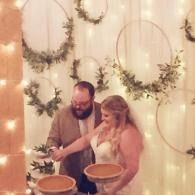 Couple cutting a wedding cake with string lights and greenery backdrop and pies on the table also