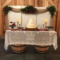 Wedding cake table with white lace cloth, candles, flowers, and rustic wooden backdrop
