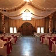 Barn-style wedding reception hall with draped white ceiling lights and red-accented tables set along both sides