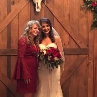 Two women posing in front of a wooden barn door, one in a white wedding dress holding a bouquet.