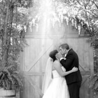 Wedding couple kissing under trees in front of a wooden gate, with sunlight streaming down