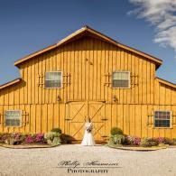 Yellow barn-style building with windows and flower beds under a blue sky, with a wedding sign in front