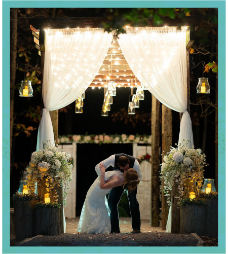 Couple kissing beneath a lit wedding arch with white curtains and floral arrangements at night