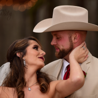 Bride and groom smiling closely, she holds his neck as he wears a beige cowboy hat and beige suit