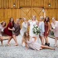 Wedding party posing in front of a wooden barn, with the bride holding a bouquet and a bridesmaid lying in front.