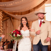 Bride in white dress and groom in beige jacket walking down a decorated wedding aisle holding a bouquet