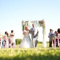 Bride and groom walking down a flower-lined wedding aisle outdoors, guests seated on both sides.