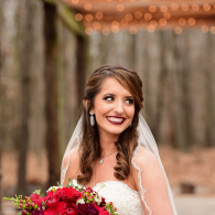 Bride in white dress and veil holding a colorful bouquet, smiling outdoors with trees and lights behind her