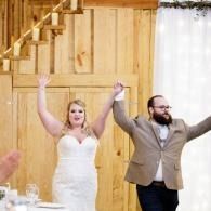 Bride and bearded groom in wedding attire raising arms in a wood-paneled reception hall