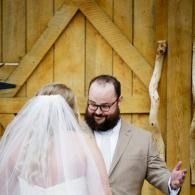 Couple at altar in a rustic wooden barn, bride in veil facing groom in tan suit, both smiling