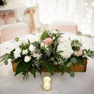 White and pink floral centerpiece in a wooden box with a small candle on a white table.