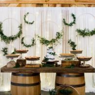 Rustic dessert table with tiered cakes, wooden barrels, and green wreaths on a white backdrop