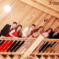 Six people leaning on a wooden railing indoors, dressed in formal attire, posing and smiling