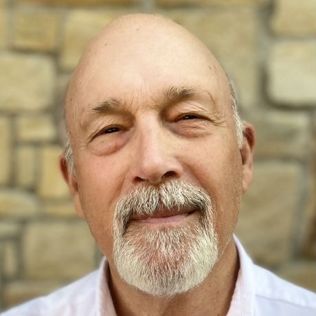 A bald man with a beard is smiling in front of a stone wall.