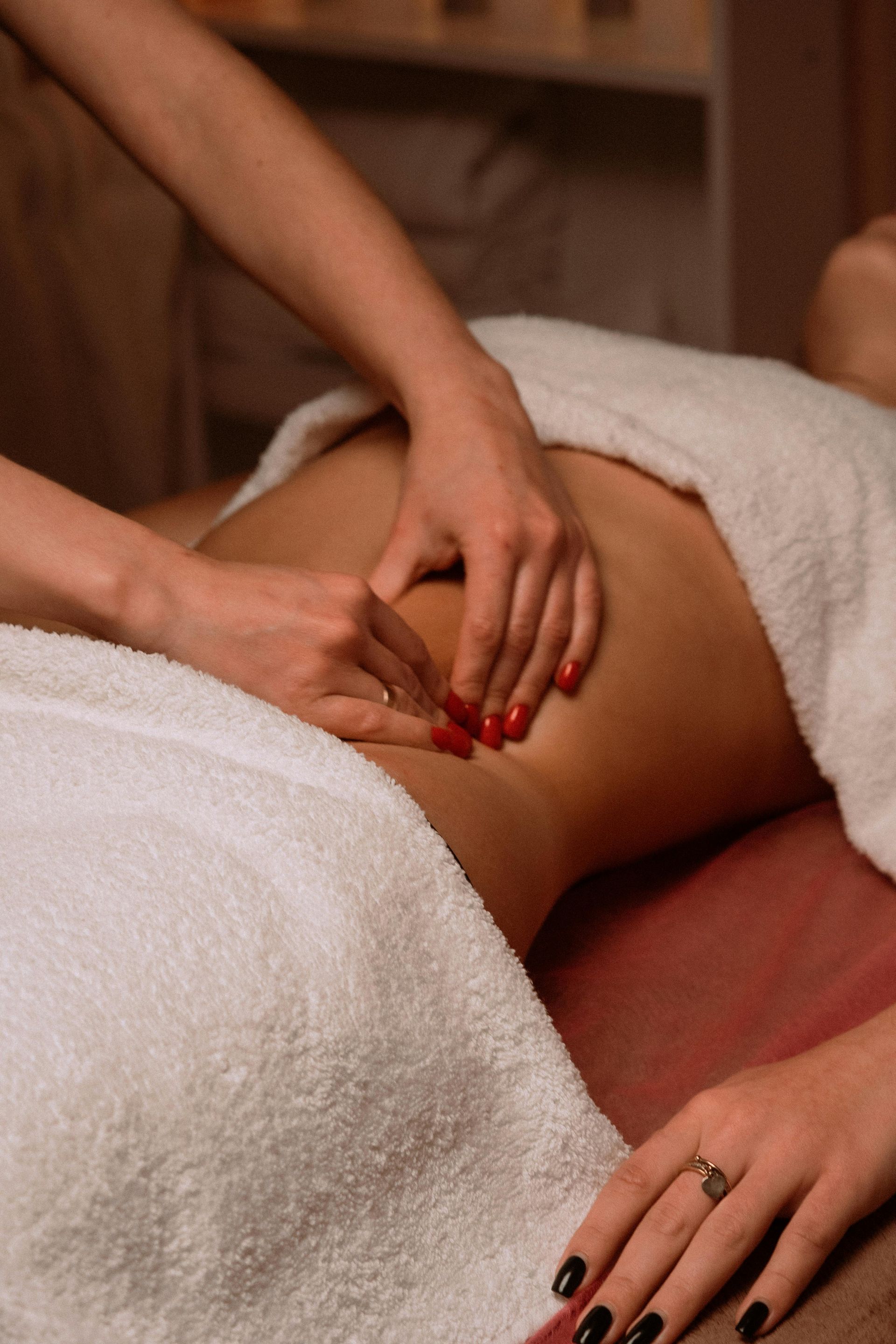 Hands massaging a person's abdomen, wrapped in a white towel, on a massage table.