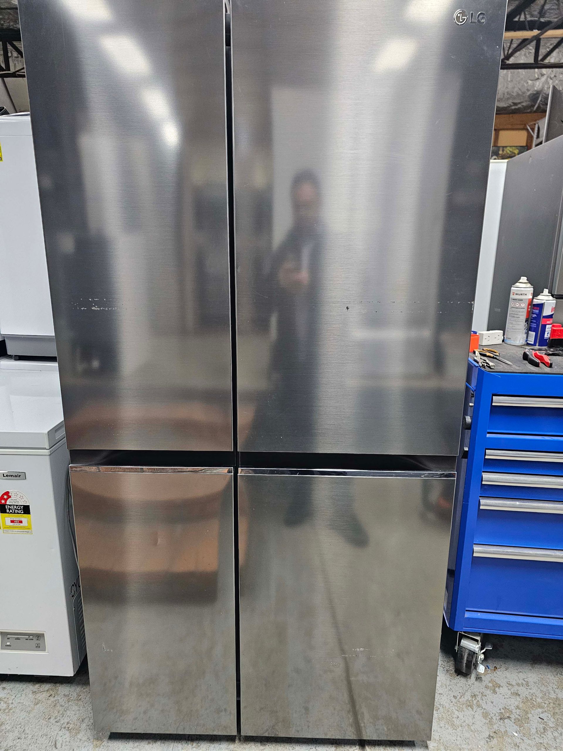 Stainless steel four-door refrigerator in a workshop, with a blue tool chest beside it.