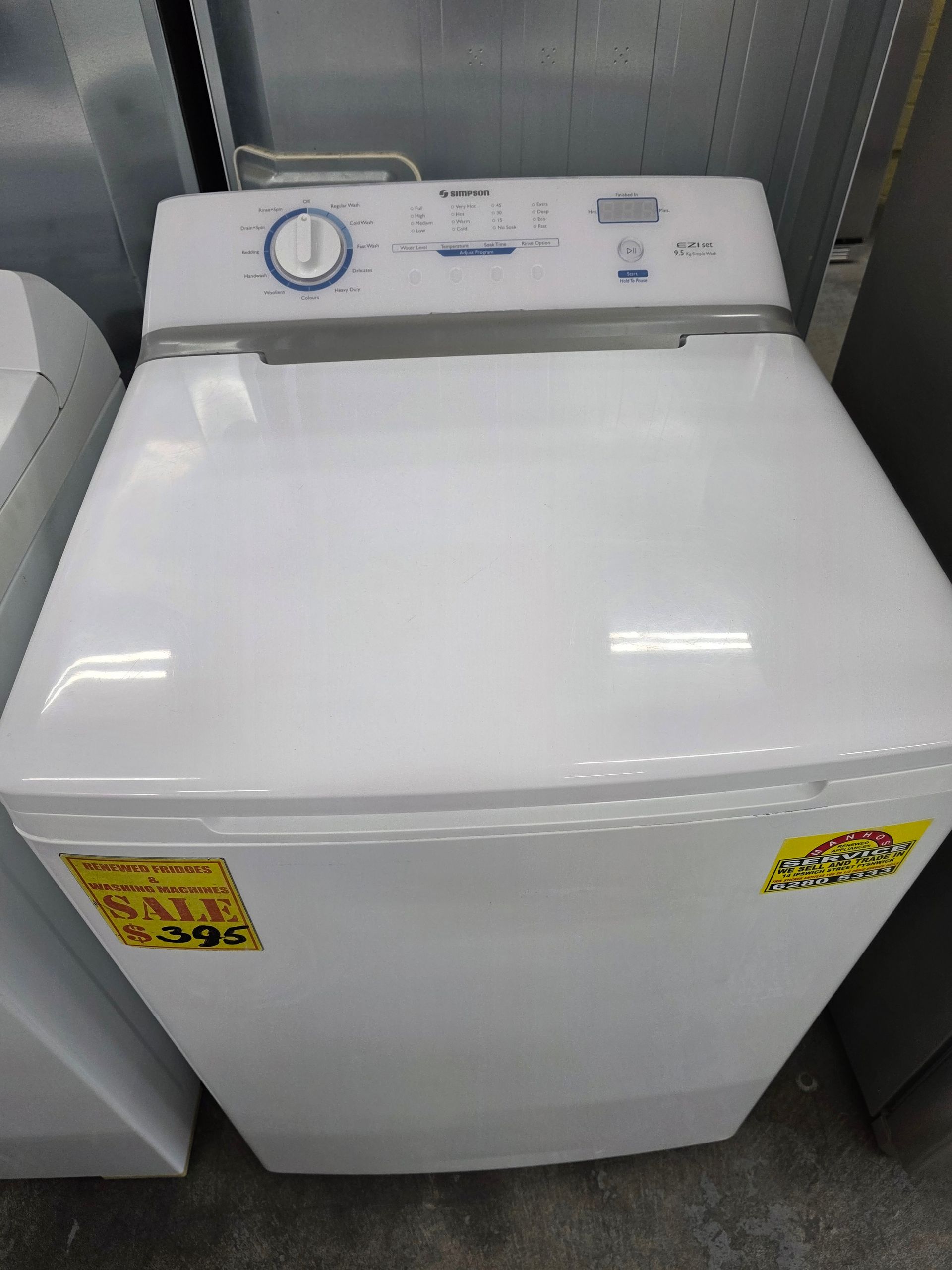 White top-load washing machine with control panel at the top. Yellow price tags on front.