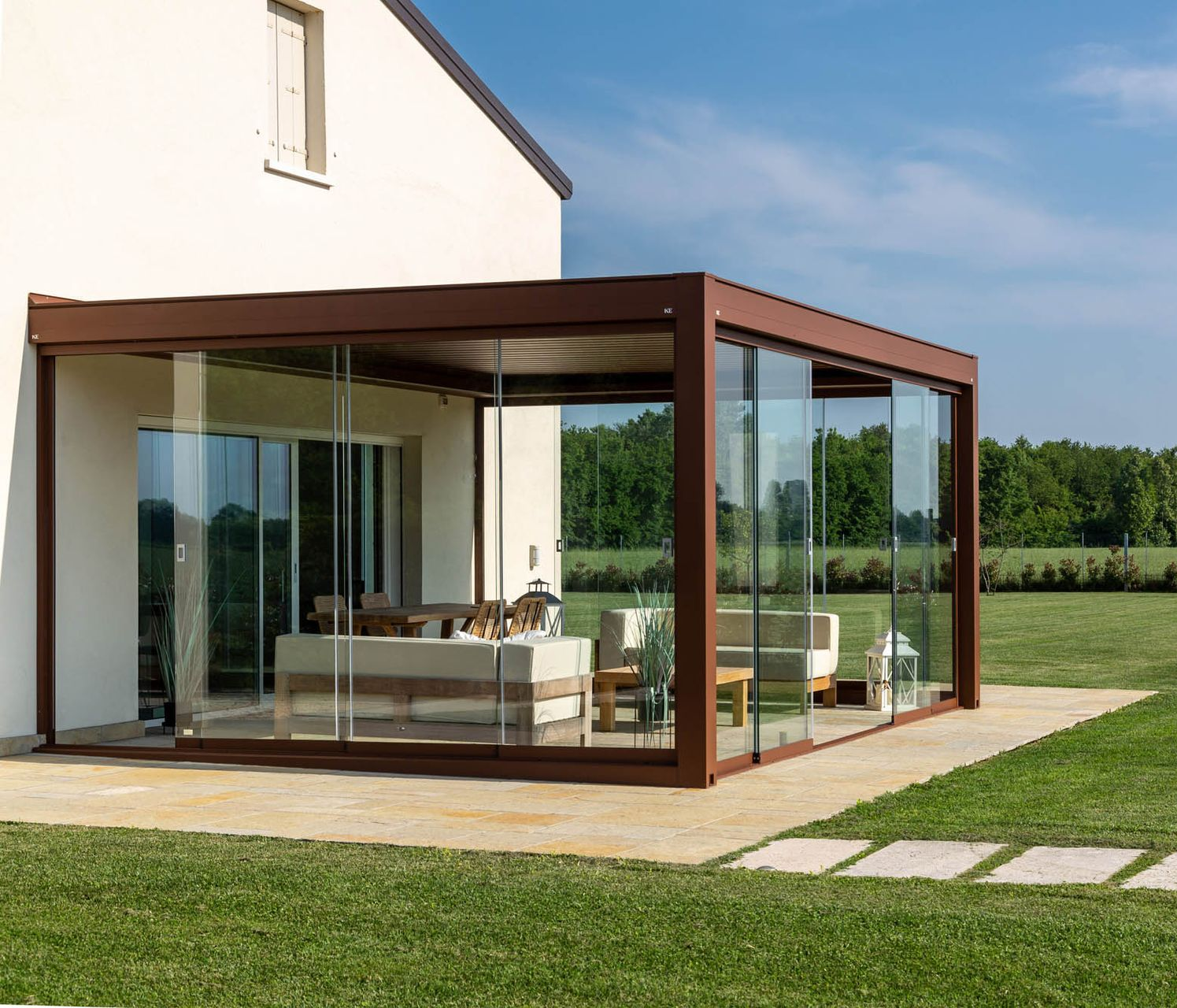 Veranda con pareti in vetro e cornice marrone, annessa a una casa bianca, con vista su un prato erboso e alberi.