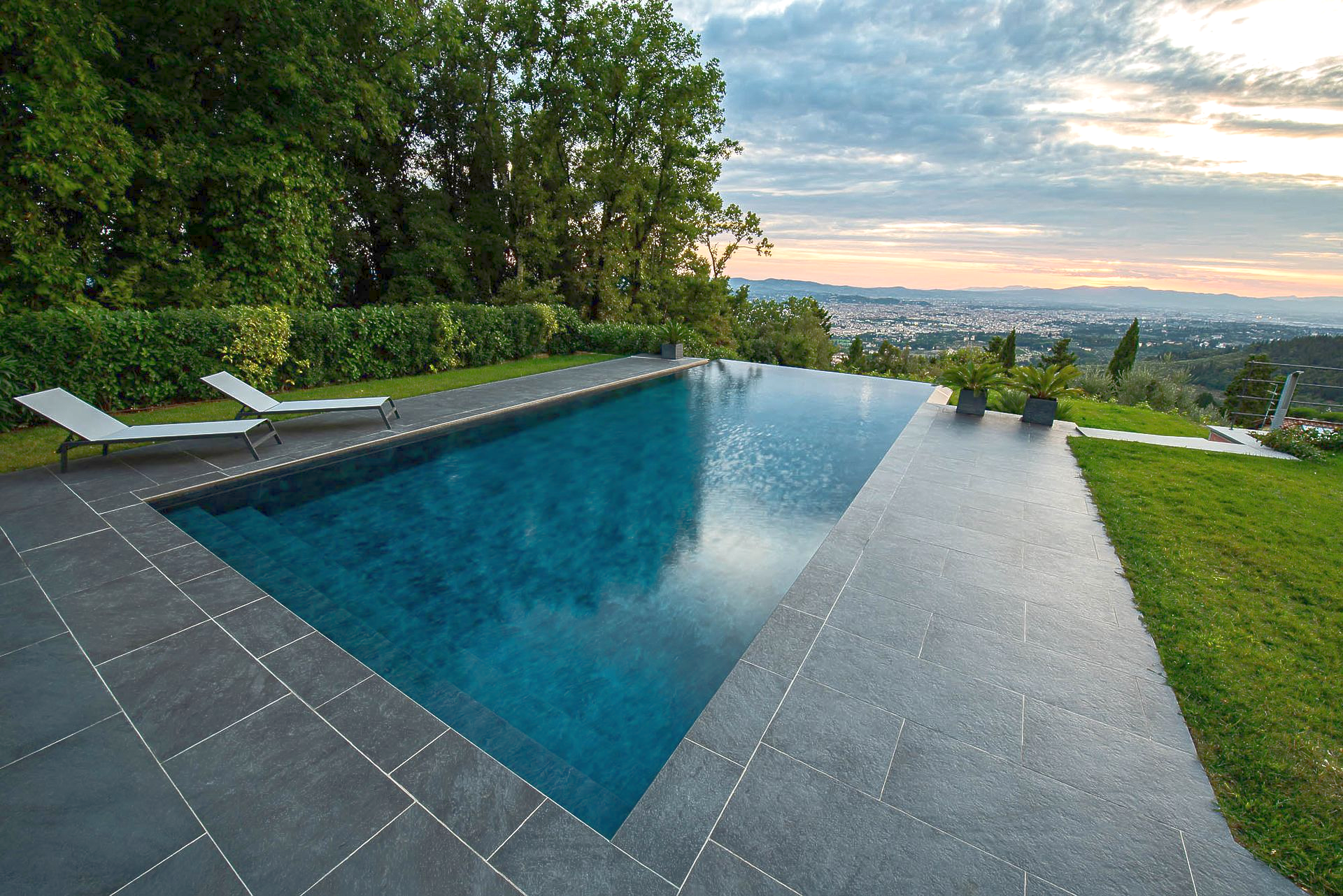 Piscina a sfioro con vista sulla città al tramonto; due sedie a sdraio sul lato.