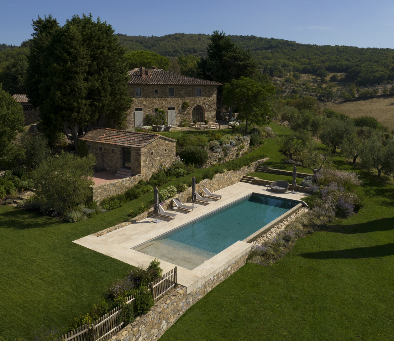 Villa in pietra con piscina immersa nel verde, con alberi e colline sullo sfondo in una giornata di sole.