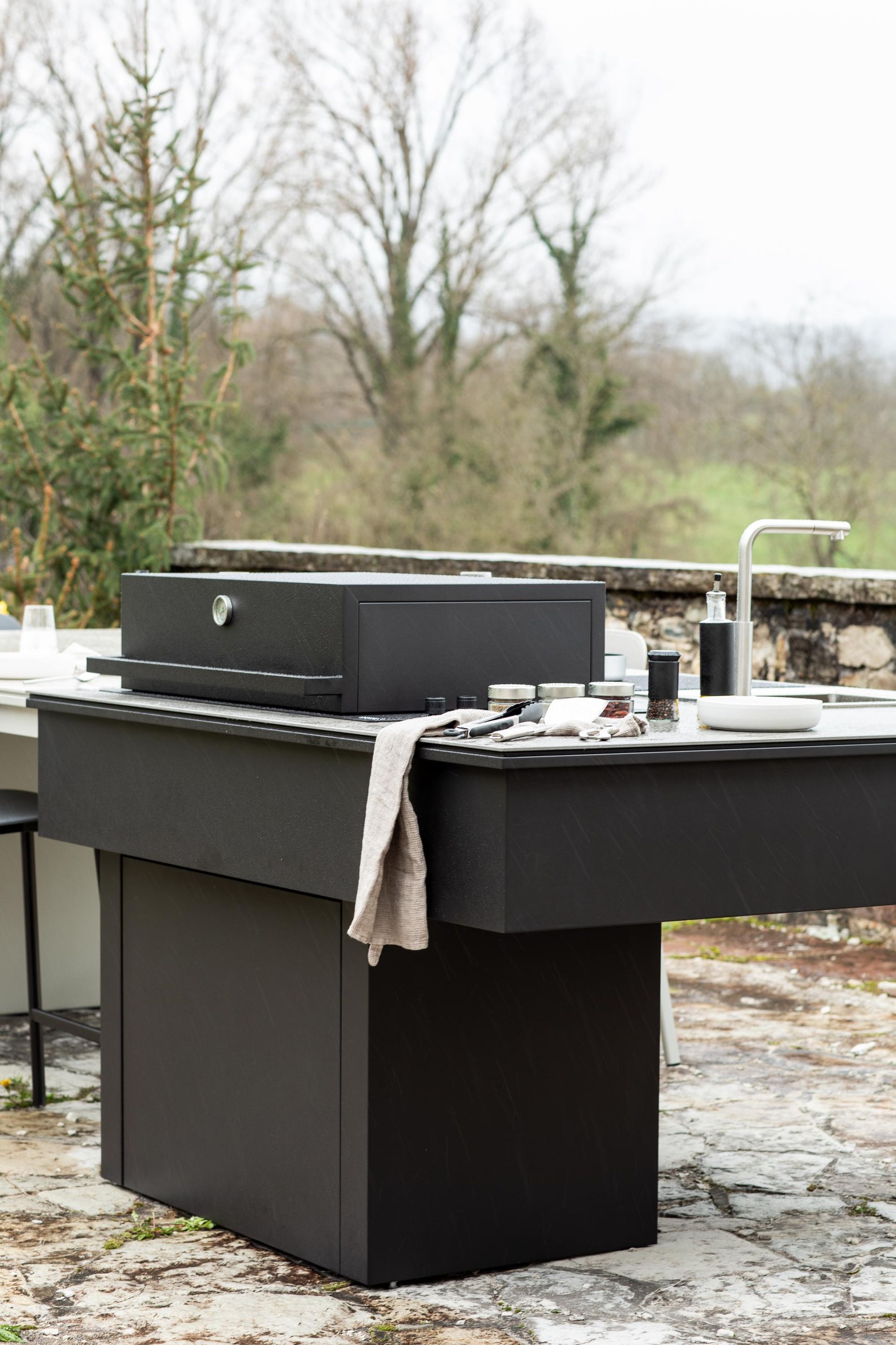 Cucina esterna nera con grill e lavello, situata su un patio in pietra con alberi sullo sfondo.