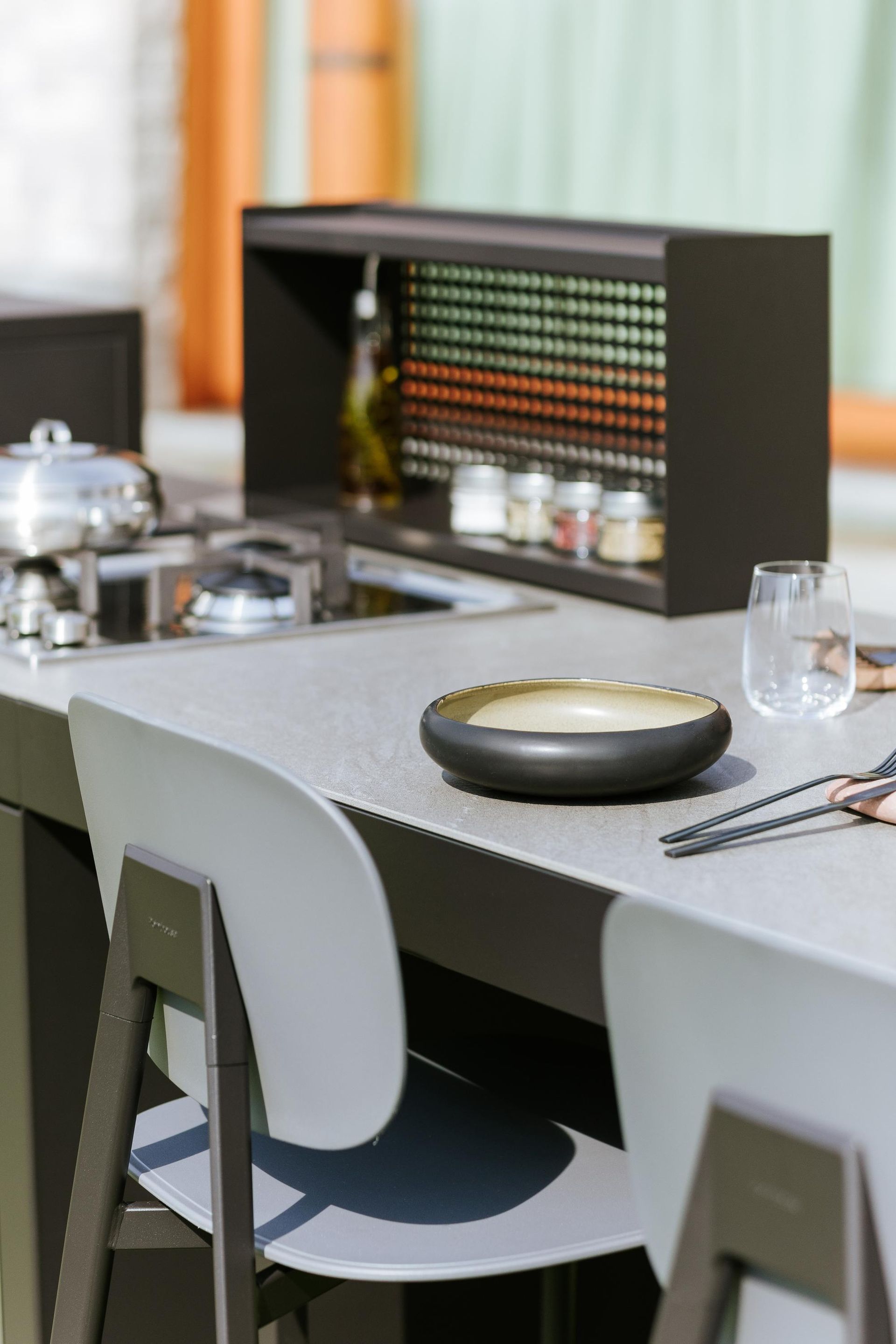 Cucina esterna con piano di lavoro grigio, fornello e porta spezie. Sedie da bar grigie.