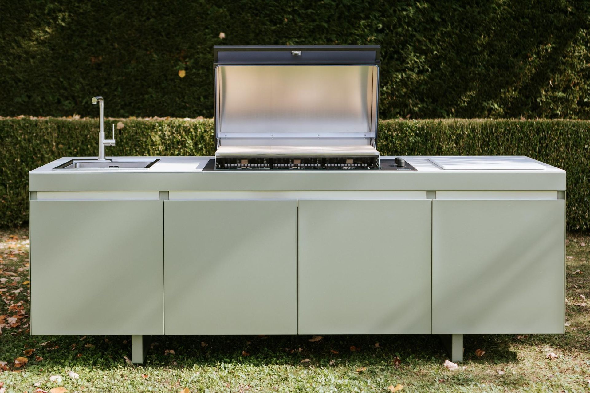 Cucina esterna con griglia in acciaio inox, lavello e mobili; situata in un cortile erboso.