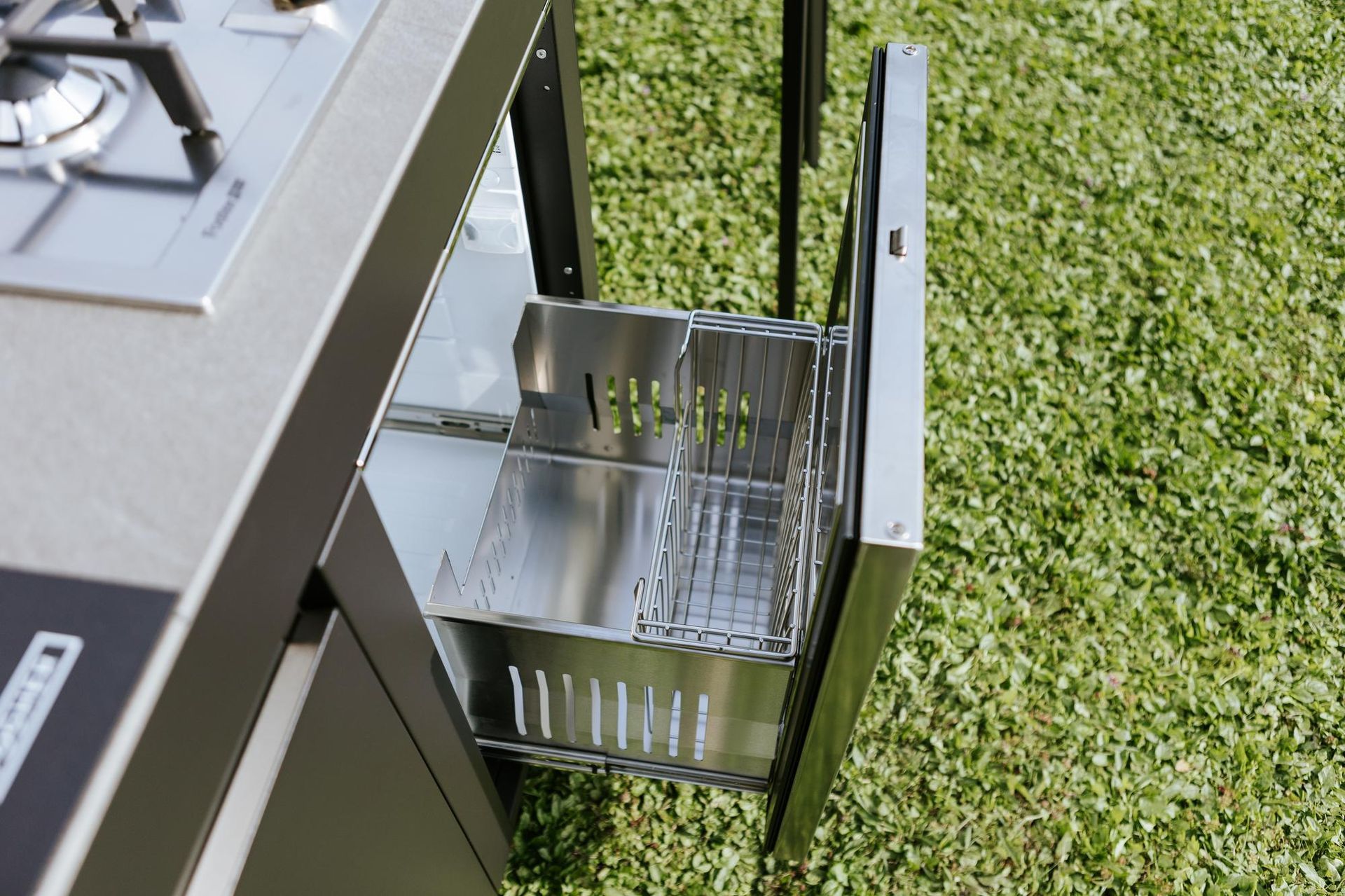 Mobile da cucina da esterno con cassetto aperto e portautensili in acciaio inox. Sfondo con erba verde.