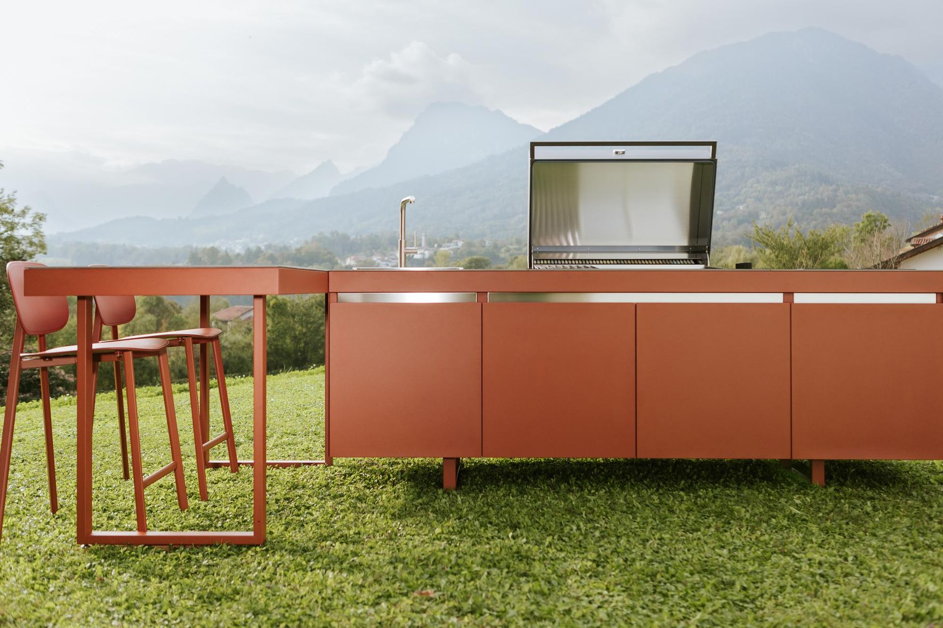 Cucina esterna con mobili color ruggine, griglia, sgabelli da bar e montagne sullo sfondo.
