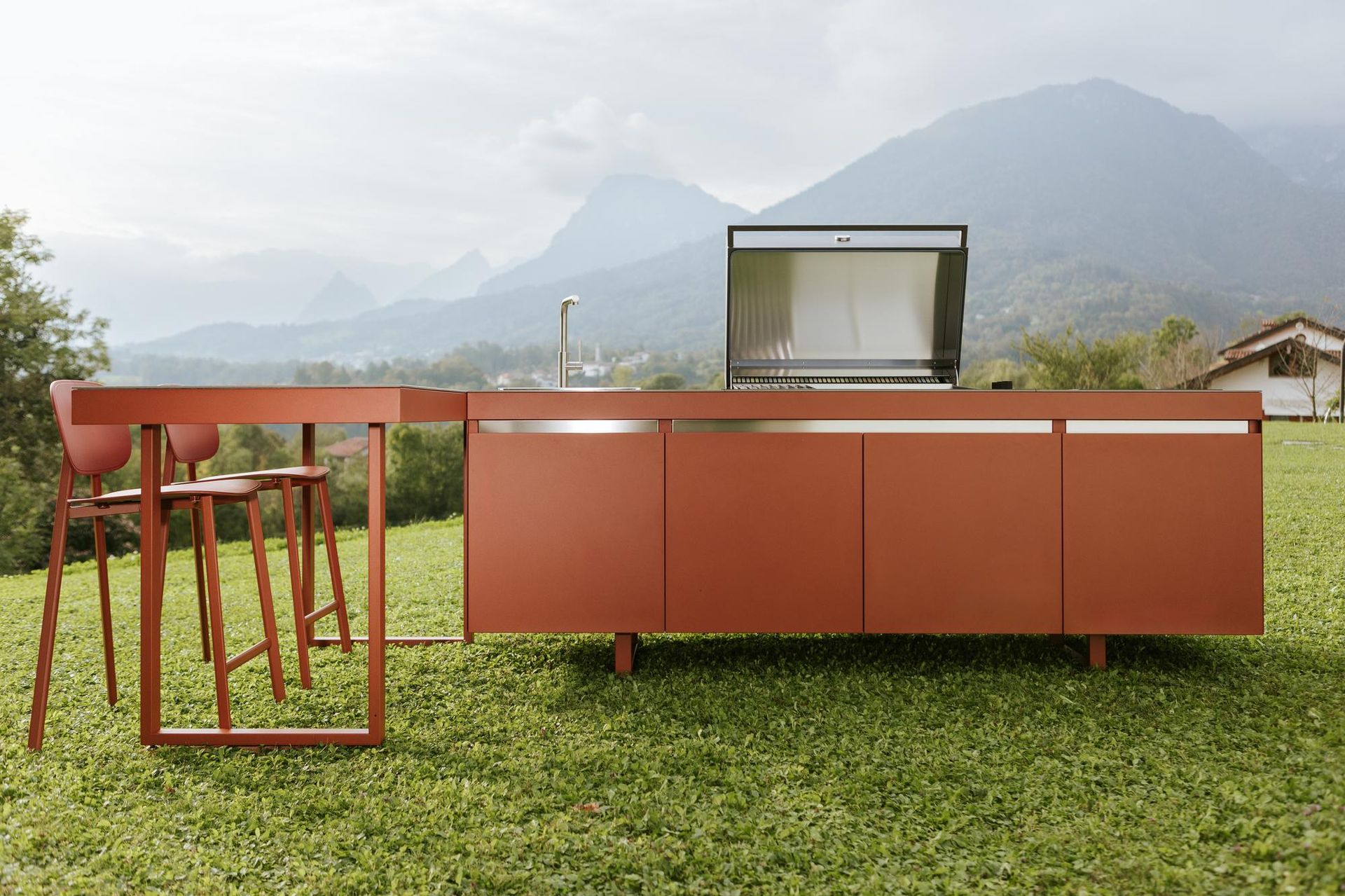 Cucina esterna con grill e posti a sedere su un prato, con le montagne sullo sfondo. Mobili color rame.