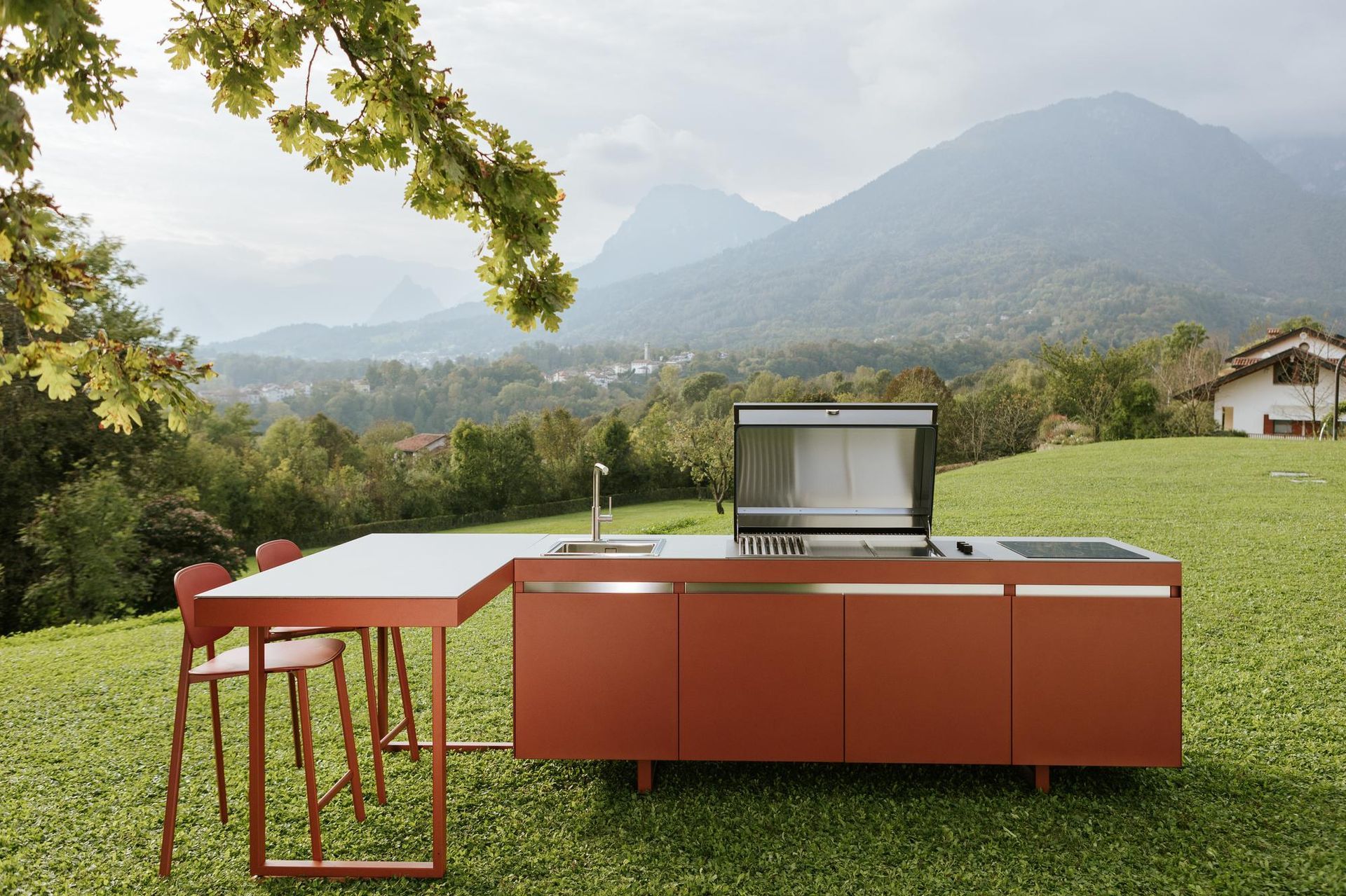 Cucina esterna con mobili rossi, griglia in acciaio inox, tavolo e sedie in un campo erboso con montagne sullo sfondo.
