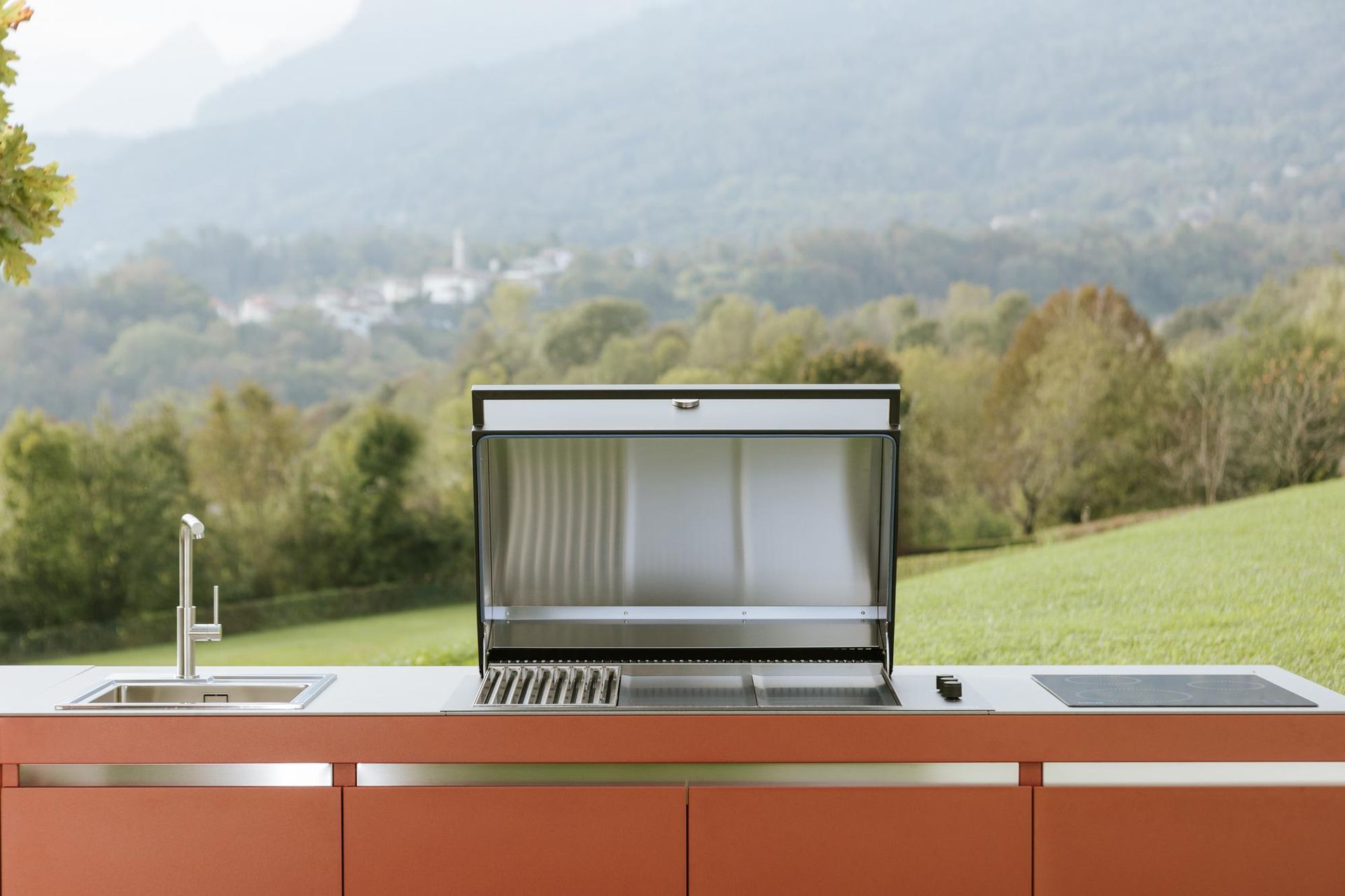 Cucina esterna con grill, lavello e piano cottura su un piano di lavoro color ruggine, adagiata su un pendio verde.