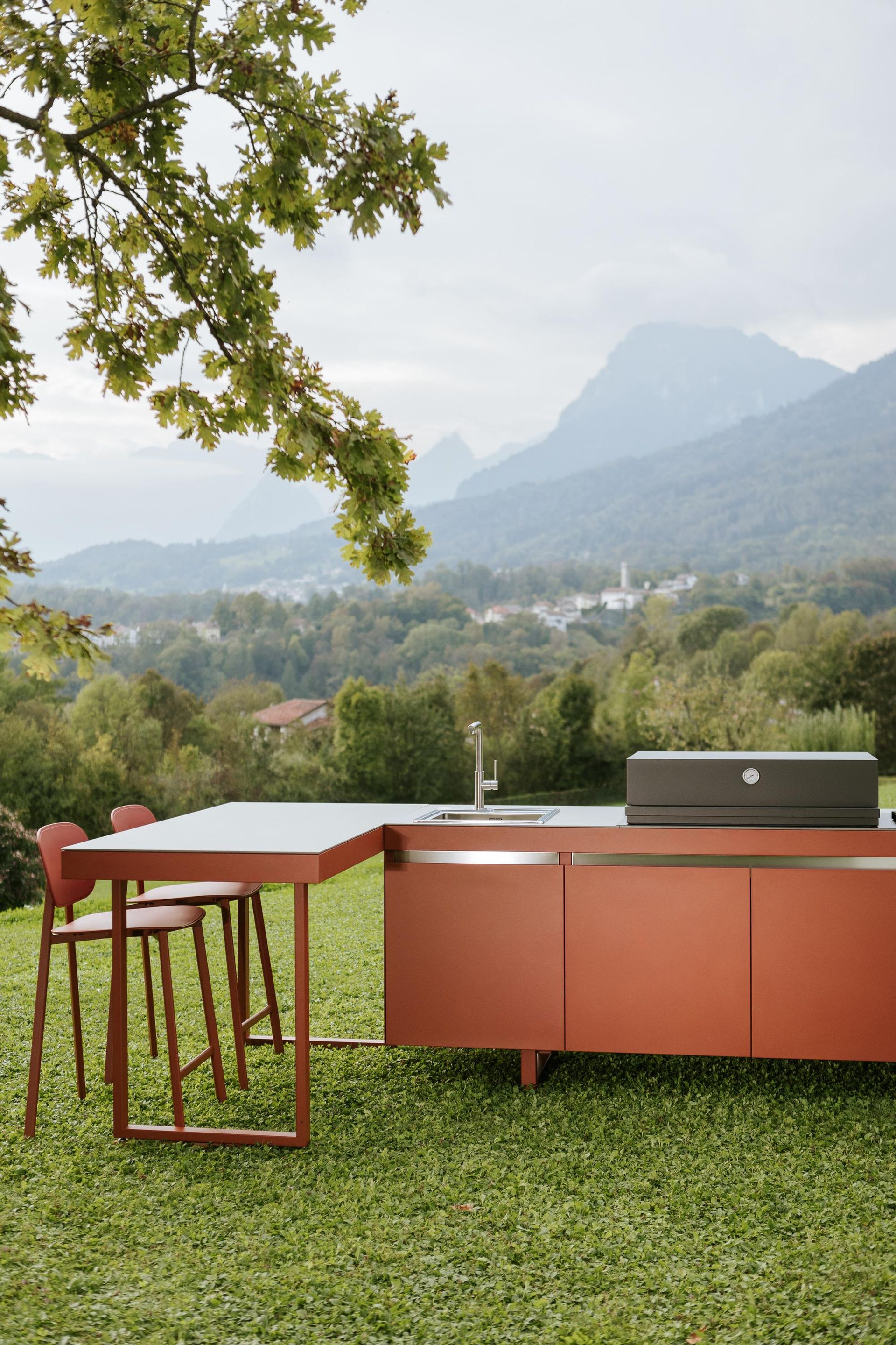 Cucina rossa all'aperto con tavolo e sgabelli su erba verde, montagne sullo sfondo.