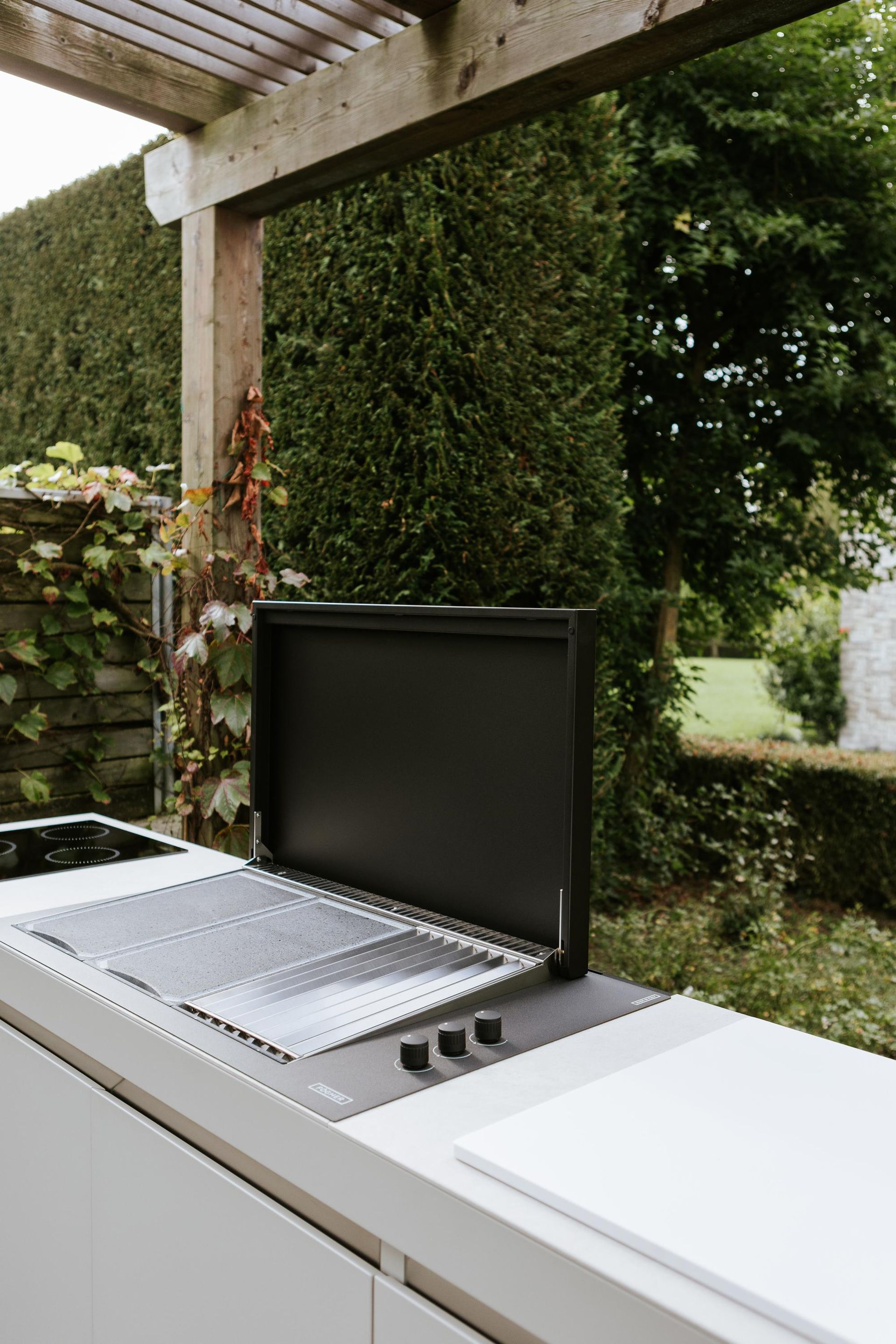 Cucina esterna con griglia a vista, immersa nel verde sotto un pergolato in legno.