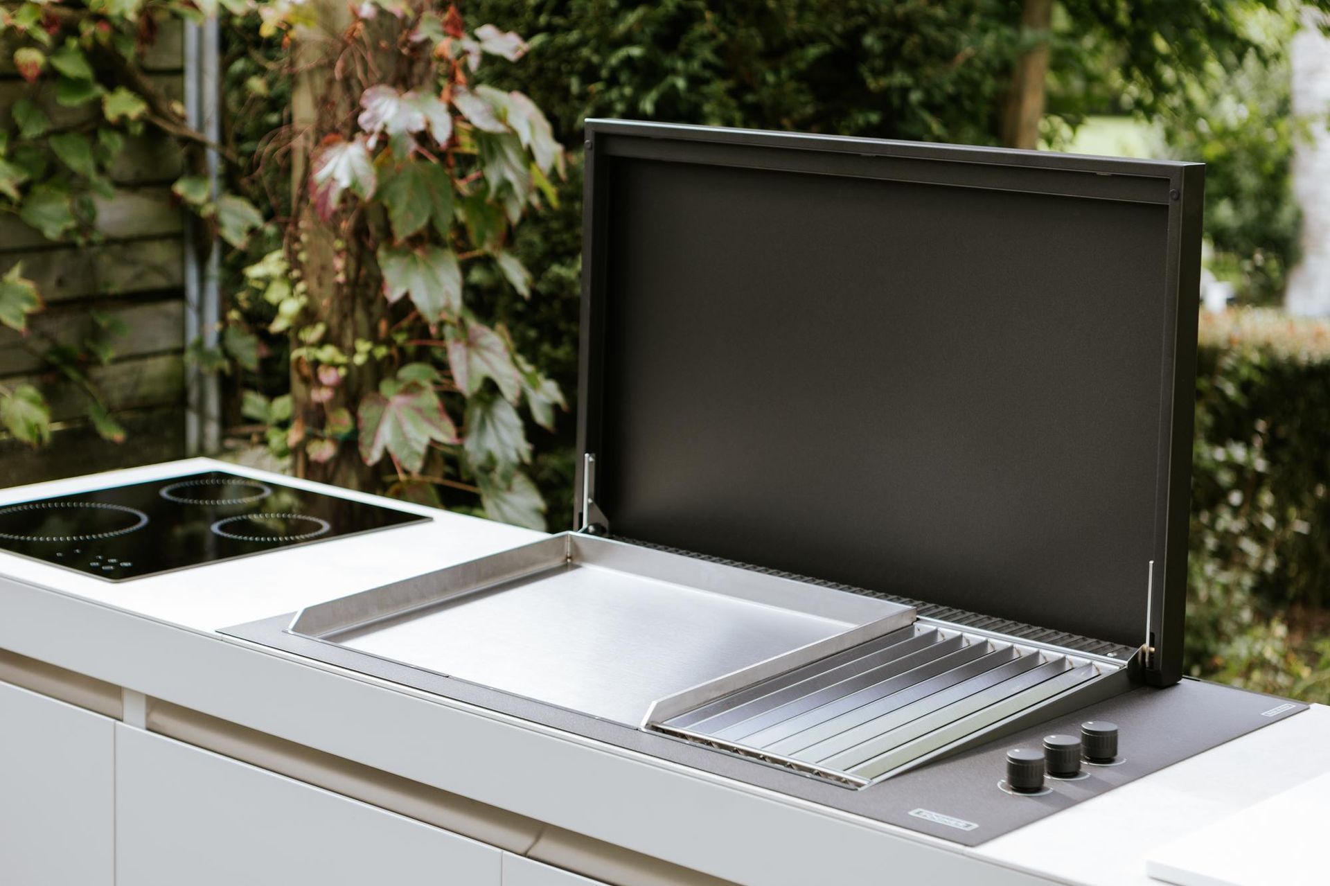 Cucina da esterno con griglia, piano cottura e coperchio aperto in un ambiente naturale.