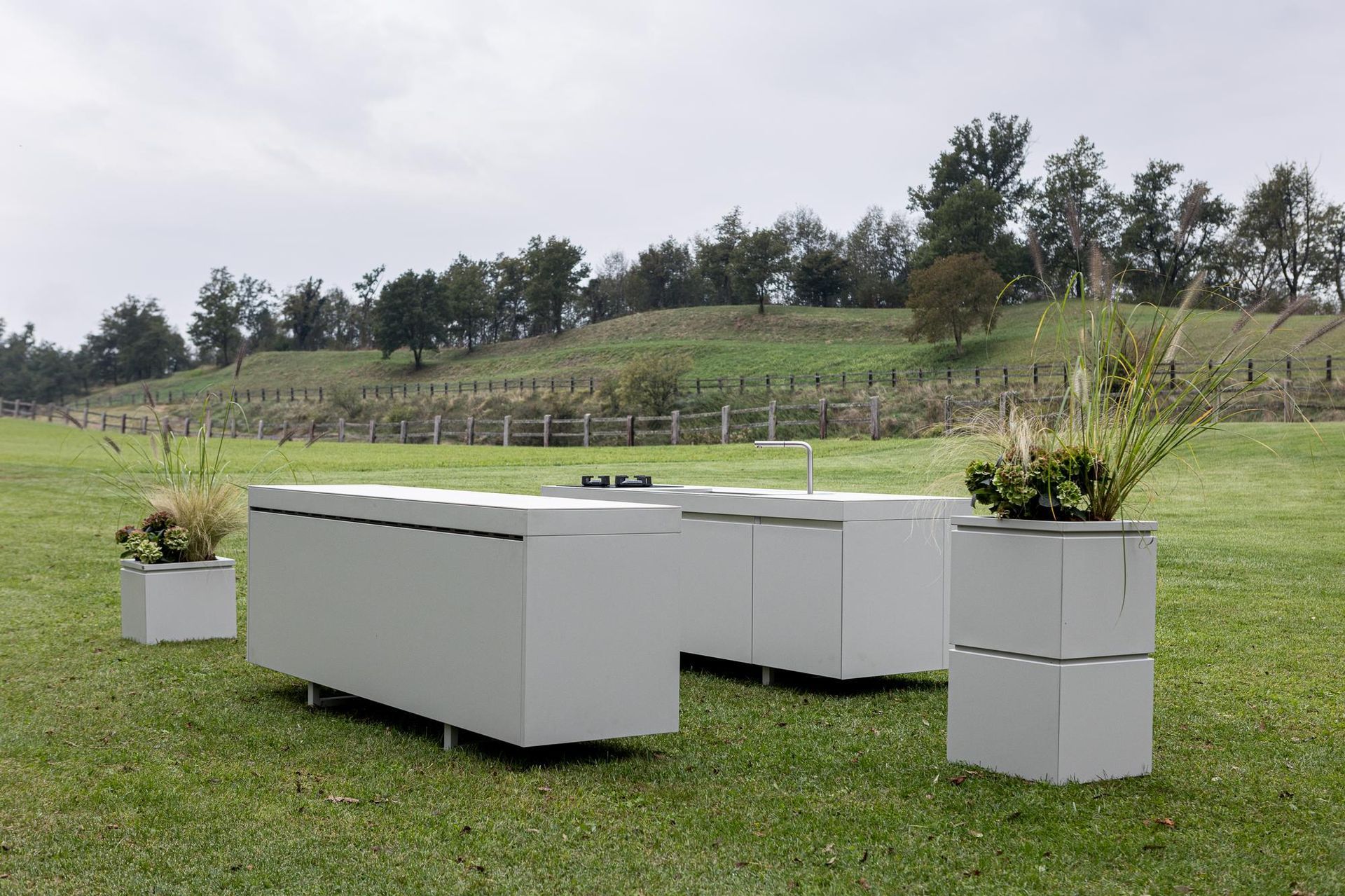 Cucina all'aperto con mobili grigi, erba e fioriere su un prato con vigneto sullo sfondo.