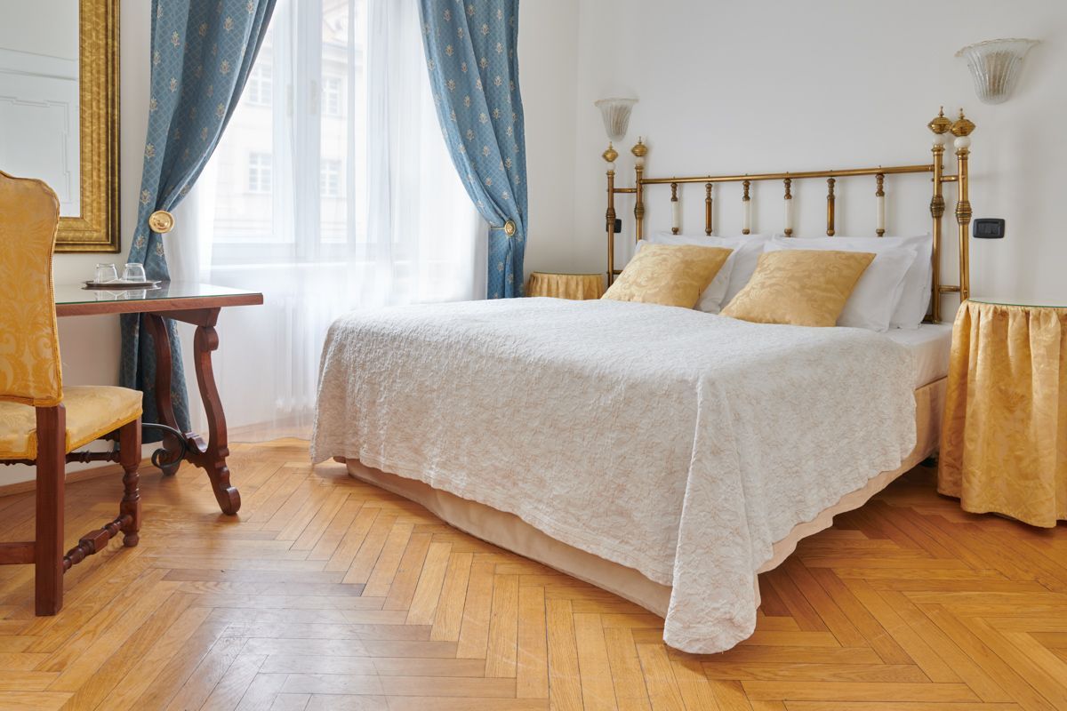 Bedroom with wooden floor, bed, window with blue curtains, small table and yellow chair.